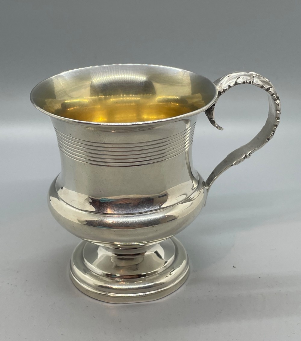 A George IV sterling silver mug featuring horizontal reeded decoration, a leaf-scrolled handle, a molded pedestal foot, and a gold-gilded interior. View II