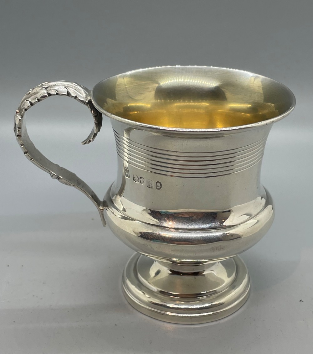 A George IV sterling silver mug featuring horizontal reeded decoration, a leaf-scrolled handle, a molded pedestal foot, and a gold-gilded interior. View IV