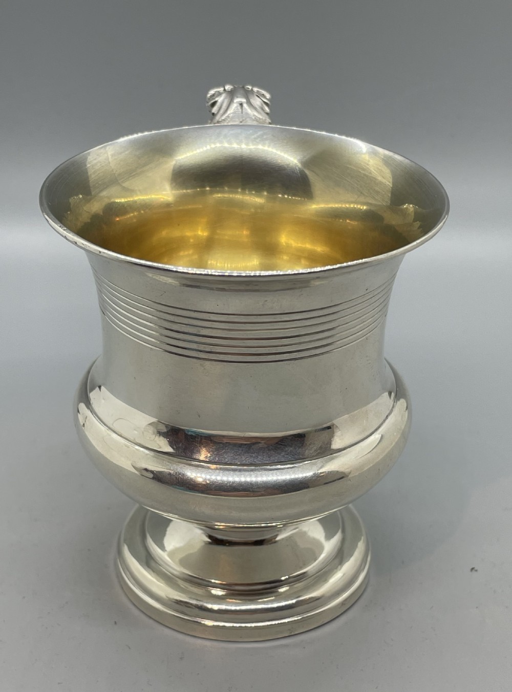 A George IV sterling silver mug featuring horizontal reeded decoration, a leaf-scrolled handle, a molded pedestal foot, and a gold-gilded interior. View V