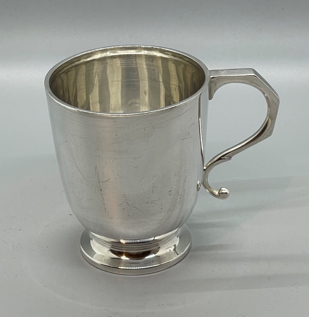 A solid sterling silver christening mug featuring a plain body, a stepped circular base, and an angular scroll handle. View II