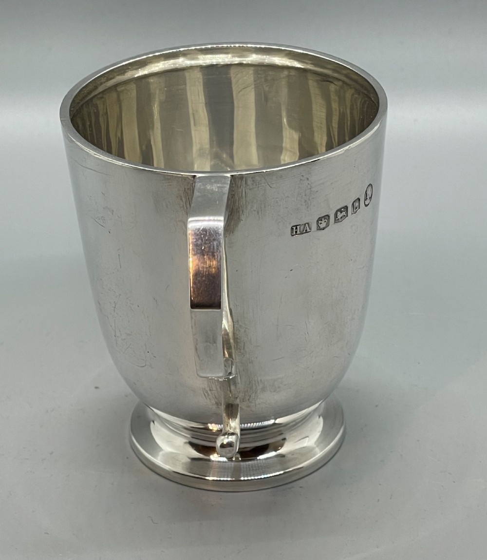 A solid sterling silver christening mug featuring a plain body, a stepped circular base, and an angular scroll handle. View III