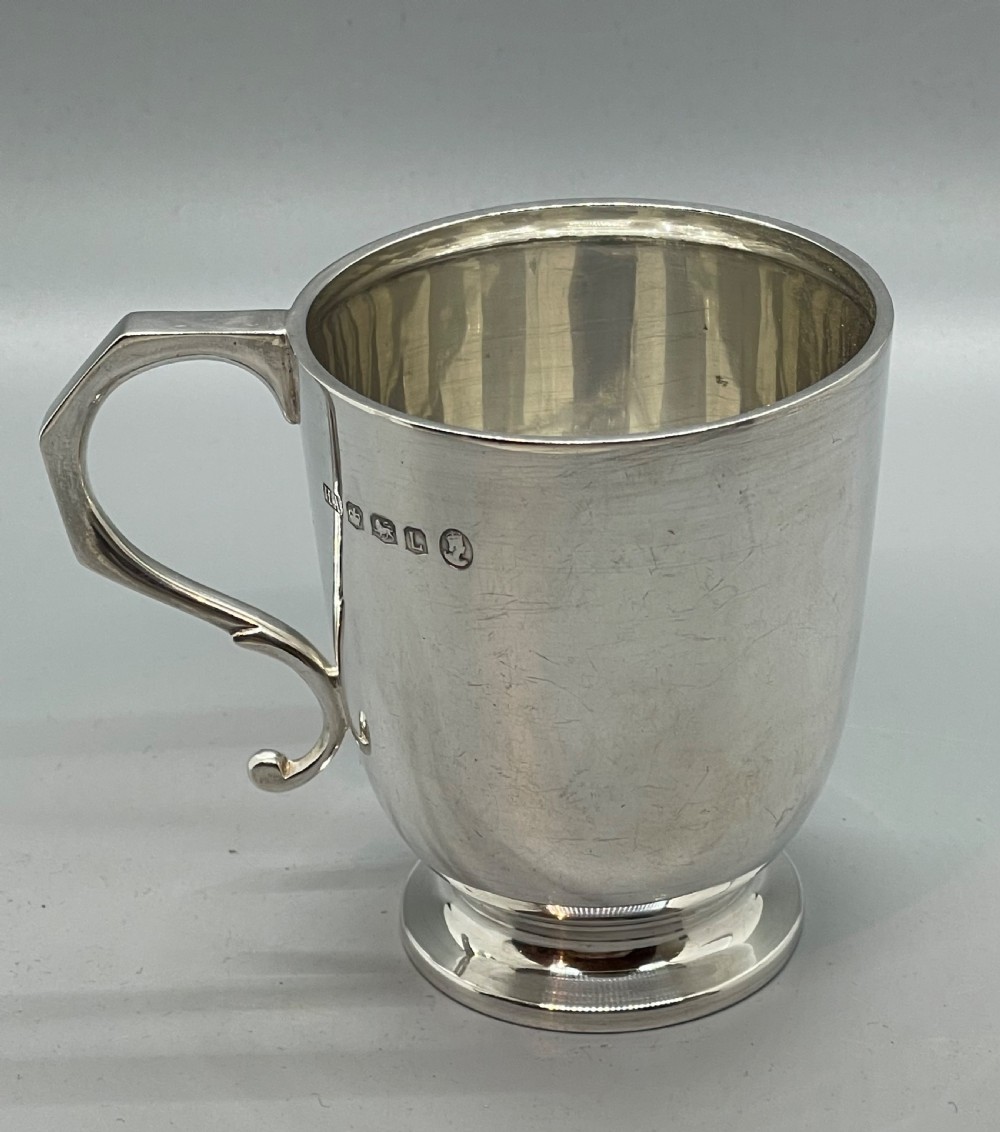 A solid sterling silver christening mug featuring a plain body, a stepped circular base, and an angular scroll handle. View IV