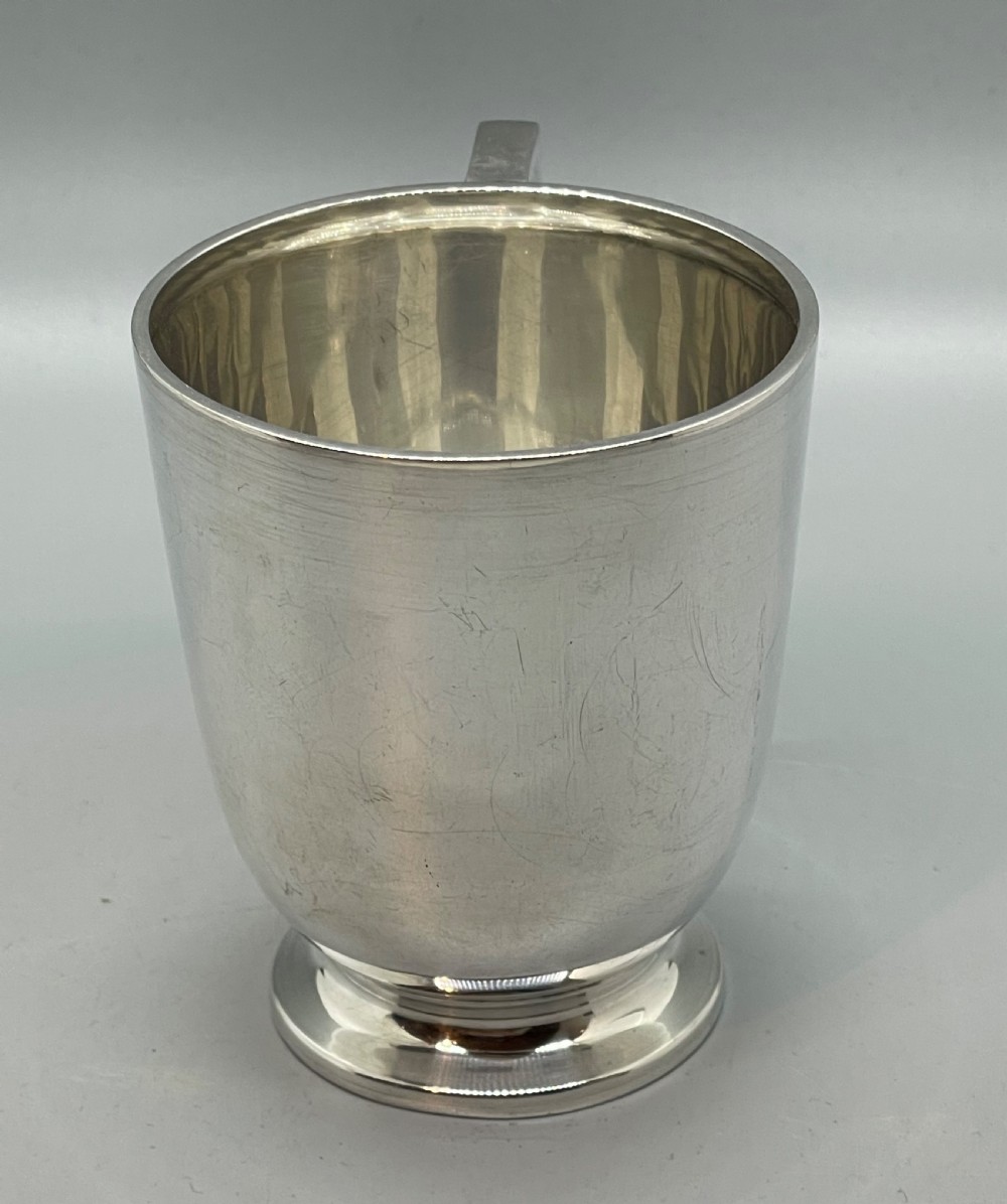 A solid sterling silver christening mug featuring a plain body, a stepped circular base, and an angular scroll handle. View V