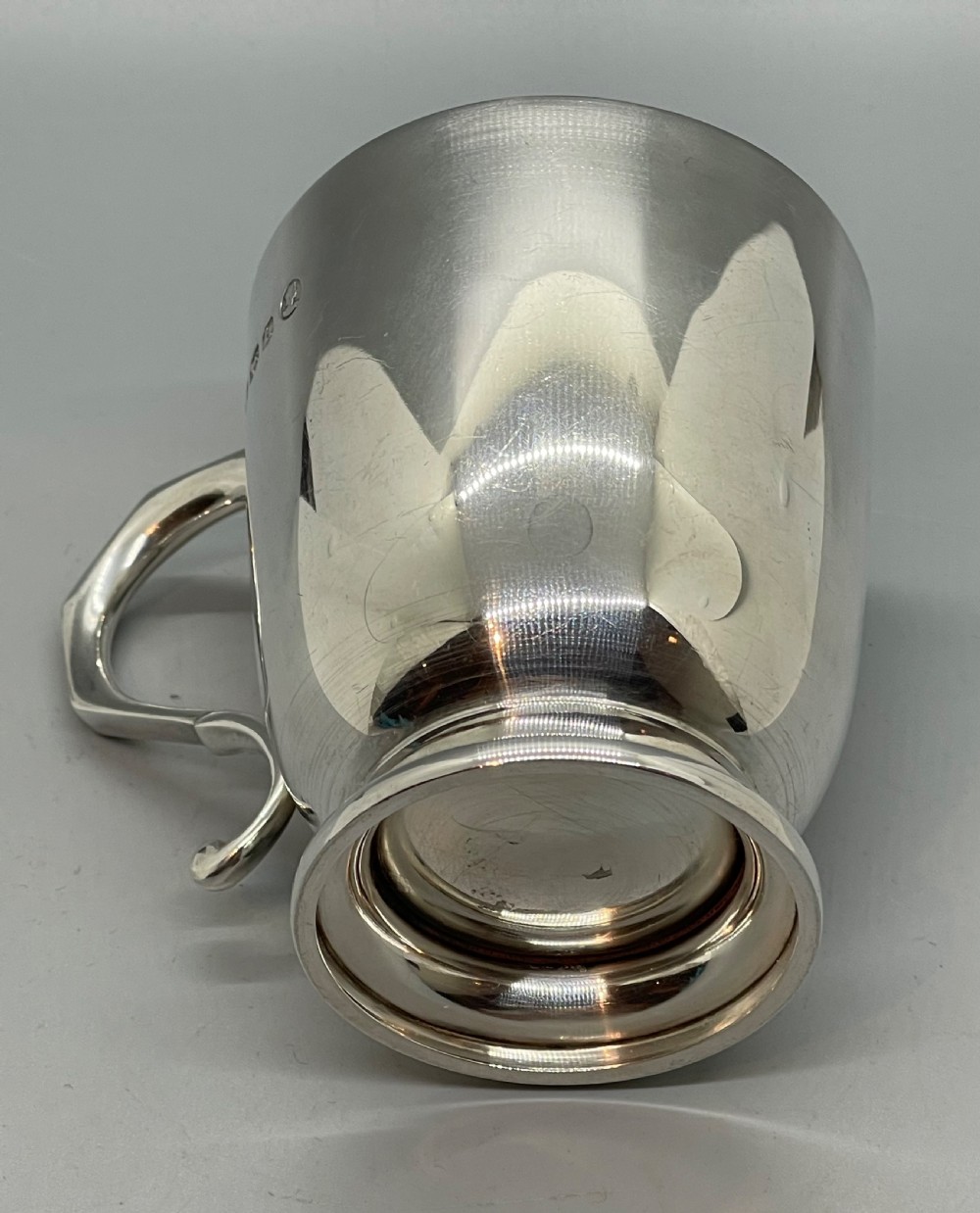 A solid sterling silver christening mug featuring a plain body, a stepped circular base, and an angular scroll handle. View VI