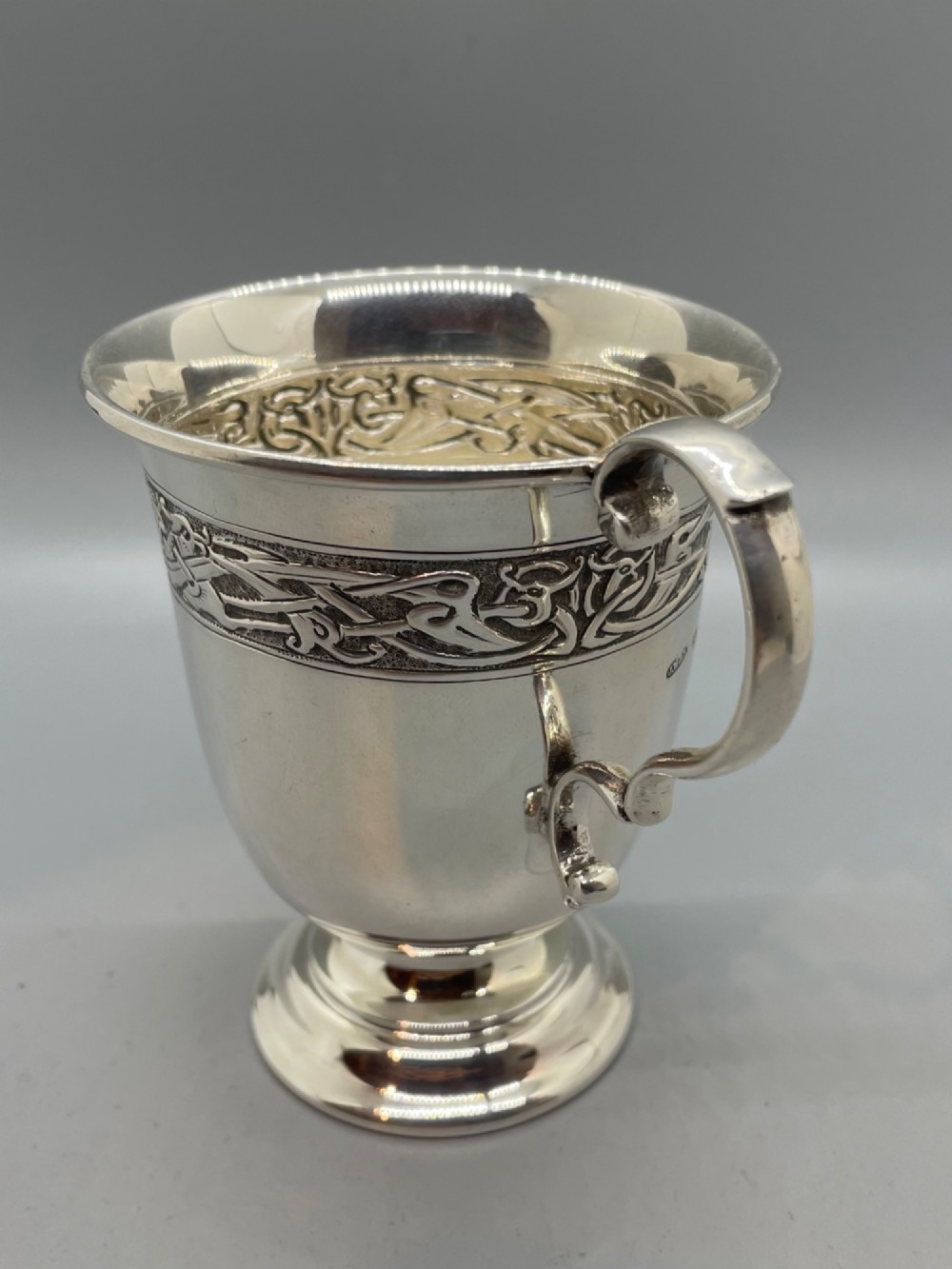 A George V sterling silver christening mug featuring a curved scroll handle, a turned pedestal foot, and an embossed Celtic knotwork band around the rim. View II