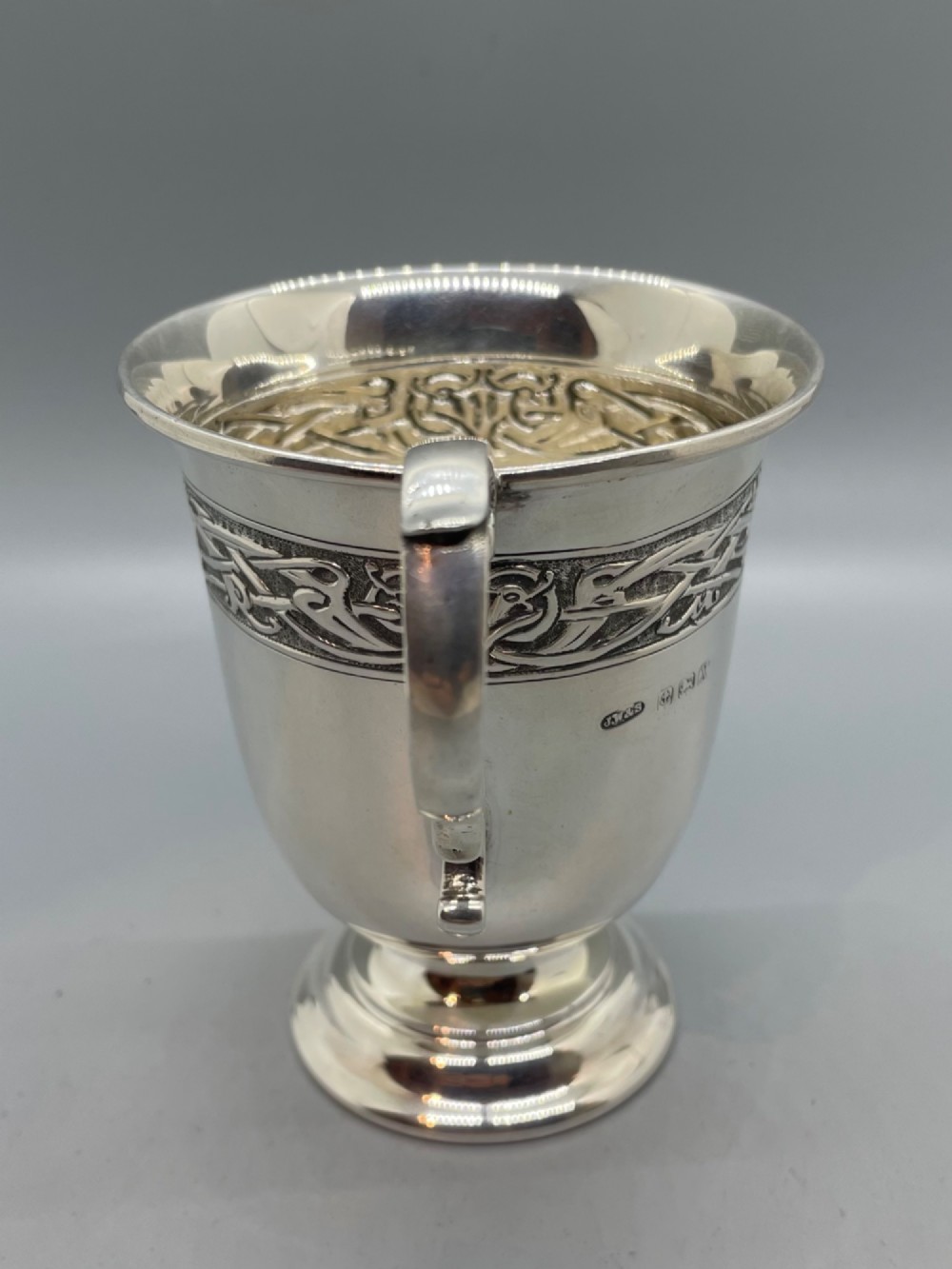 A George V sterling silver christening mug featuring a curved scroll handle, a turned pedestal foot, and an embossed Celtic knotwork band around the rim. View III