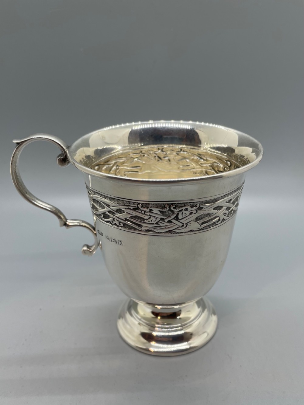A George V sterling silver christening mug featuring a curved scroll handle, a turned pedestal foot, and an embossed Celtic knotwork band around the rim. View IV
