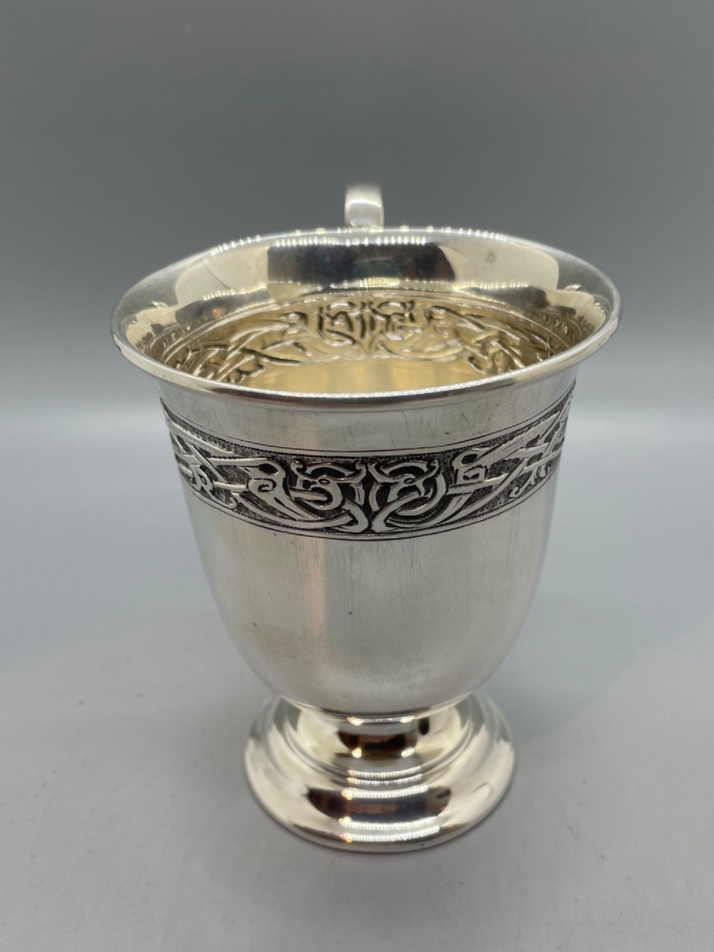 A George V sterling silver christening mug featuring a curved scroll handle, a turned pedestal foot, and an embossed Celtic knotwork band around the rim. View V