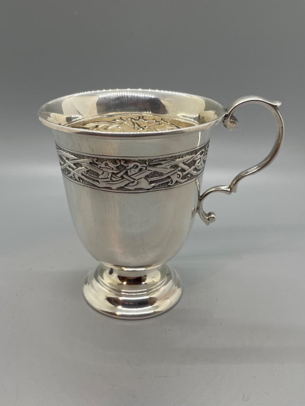 A George V sterling silver christening mug featuring a curved scroll handle, a turned pedestal foot, and an embossed Celtic knotwork band around the rim. View VI