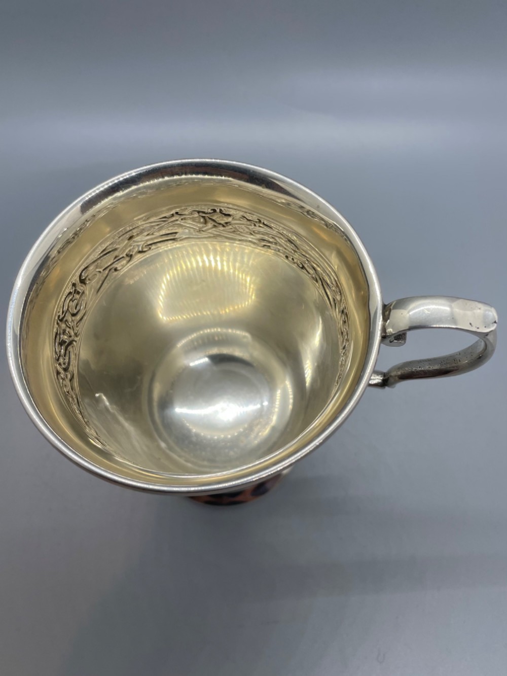 A George V sterling silver christening mug featuring a curved scroll handle, a turned pedestal foot, and an embossed Celtic knotwork band around the rim. View VII