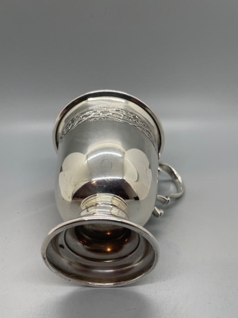 A George V sterling silver christening mug featuring a curved scroll handle, a turned pedestal foot, and an embossed Celtic knotwork band around the rim. View VIII