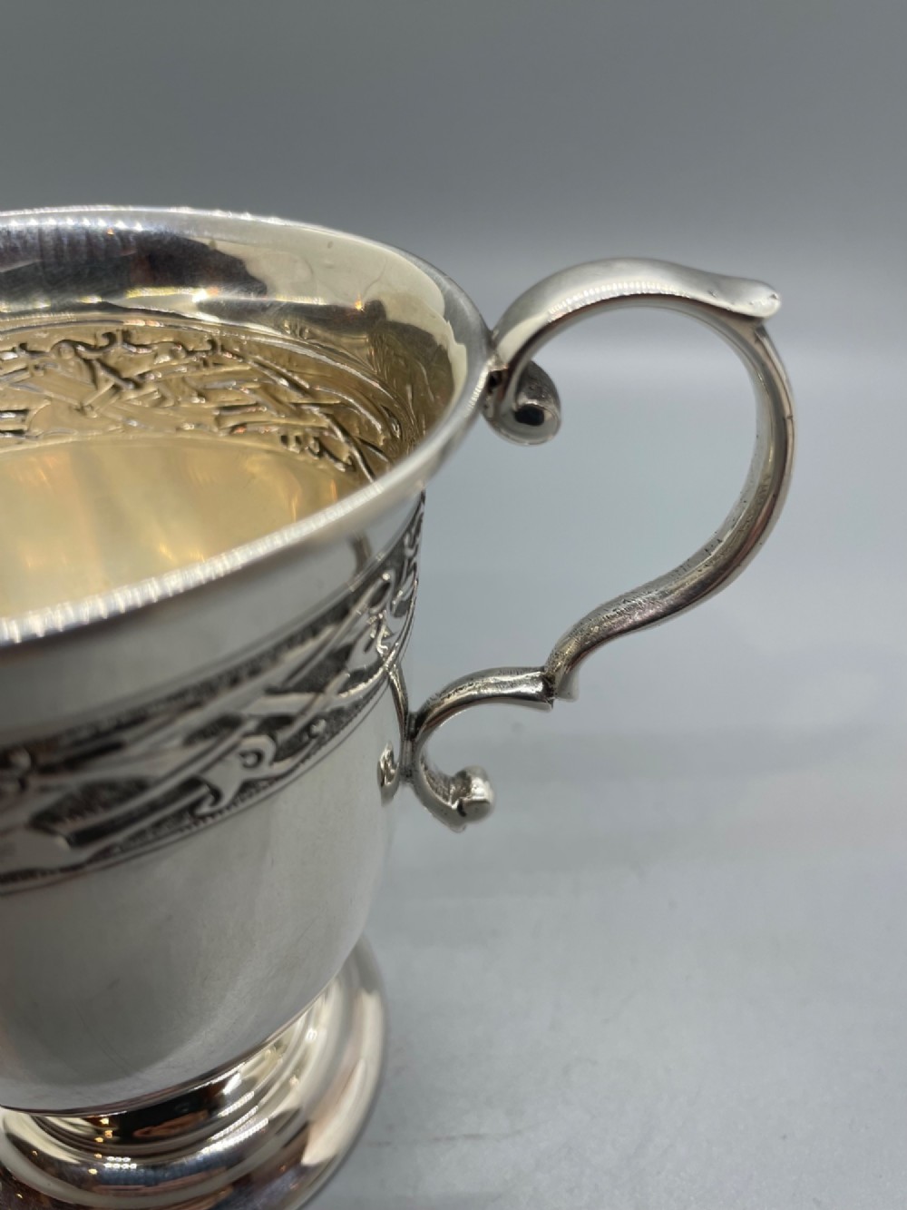 A George V sterling silver christening mug featuring a curved scroll handle, a turned pedestal foot, and an embossed Celtic knotwork band around the rim. View IX