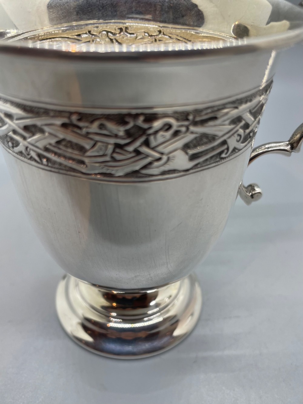 A George V sterling silver christening mug featuring a curved scroll handle, a turned pedestal foot, and an embossed Celtic knotwork band around the rim. View X