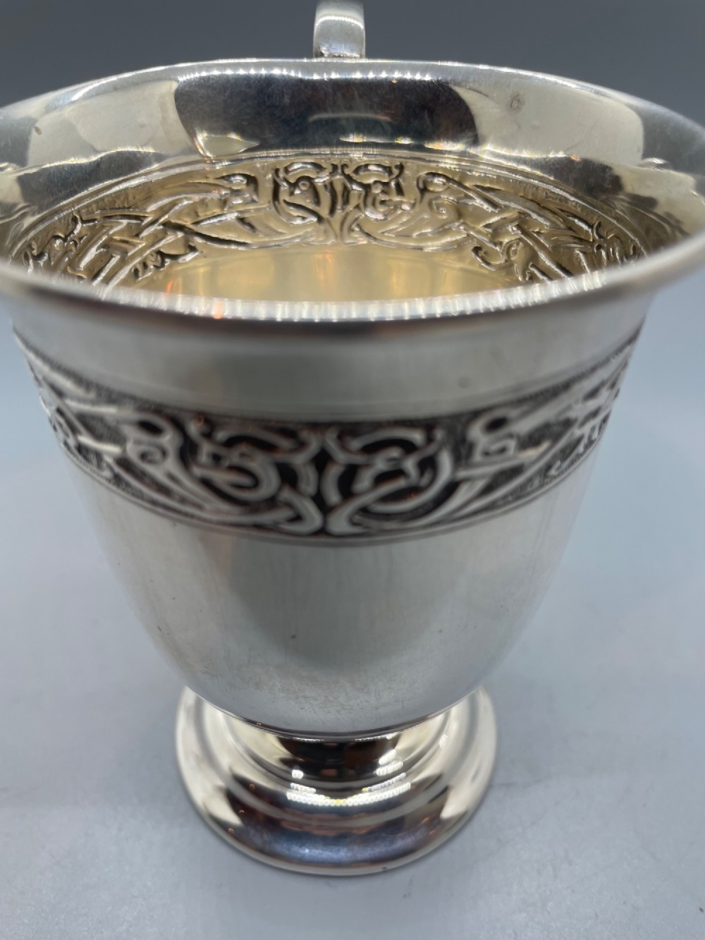 A George V sterling silver christening mug featuring a curved scroll handle, a turned pedestal foot, and an embossed Celtic knotwork band around the rim. View XI