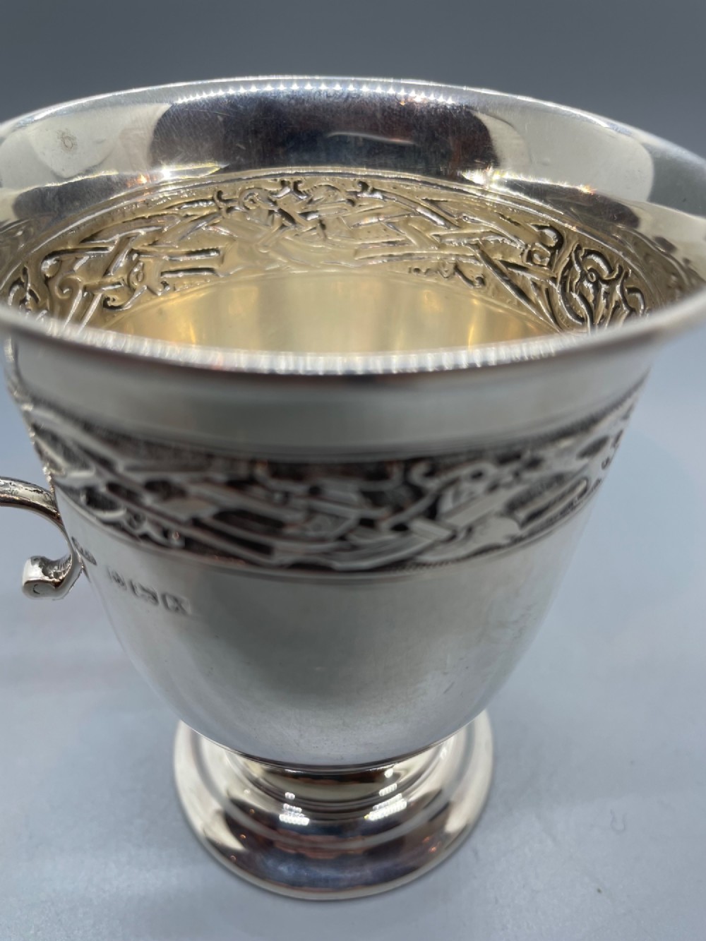 A George V sterling silver christening mug featuring a curved scroll handle, a turned pedestal foot, and an embossed Celtic knotwork band around the rim. View XII