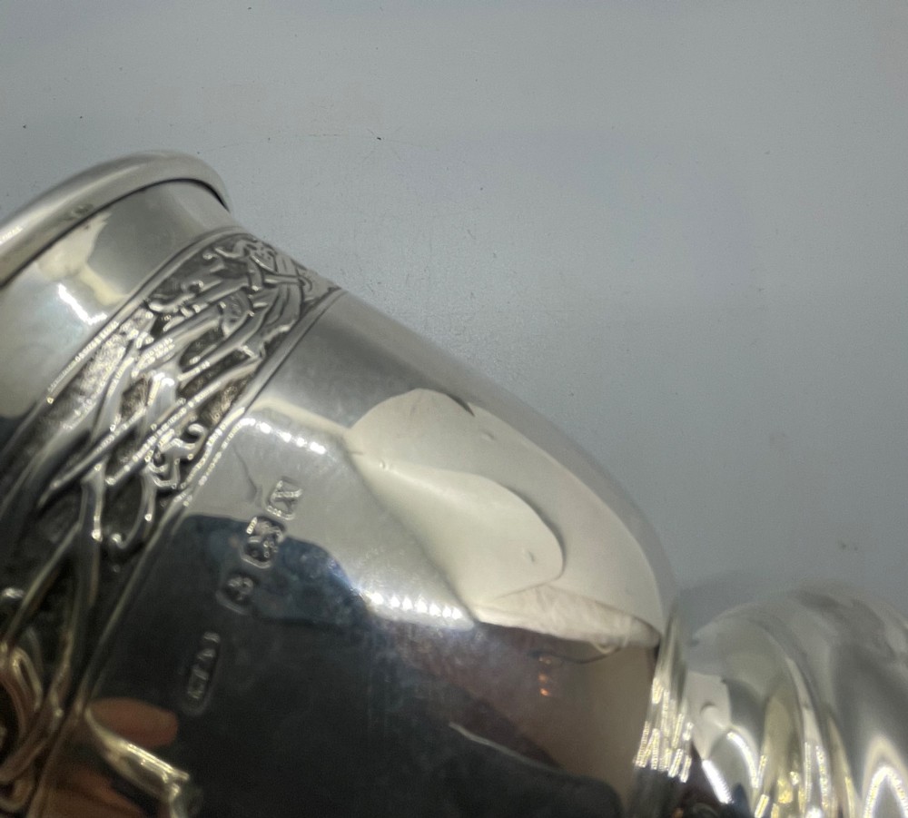 A George V sterling silver christening mug featuring a curved scroll handle, a turned pedestal foot, and an embossed Celtic knotwork band around the rim. View XIII