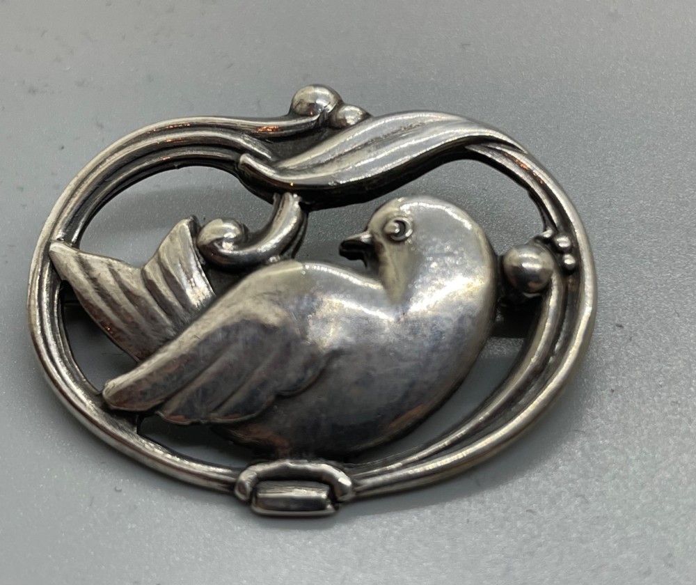 An oval sterling silver brooch depicting a bird in relief surrounded by stylised leaves and silver beads.