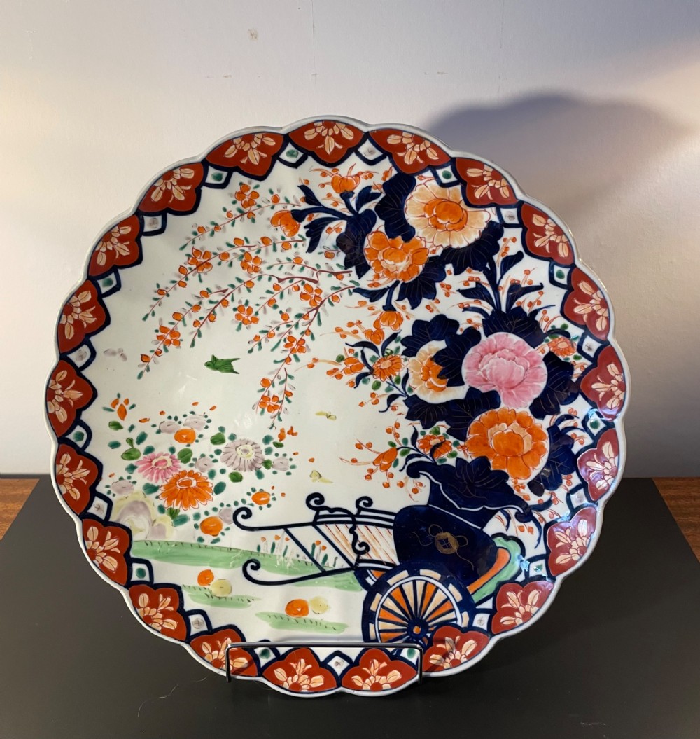 A circular Japanese Imari porcelain charger with a scalloped rim, featuring a hand-painted floral cart motif surrounded by cobalt blue and rust red enamel borders. View II