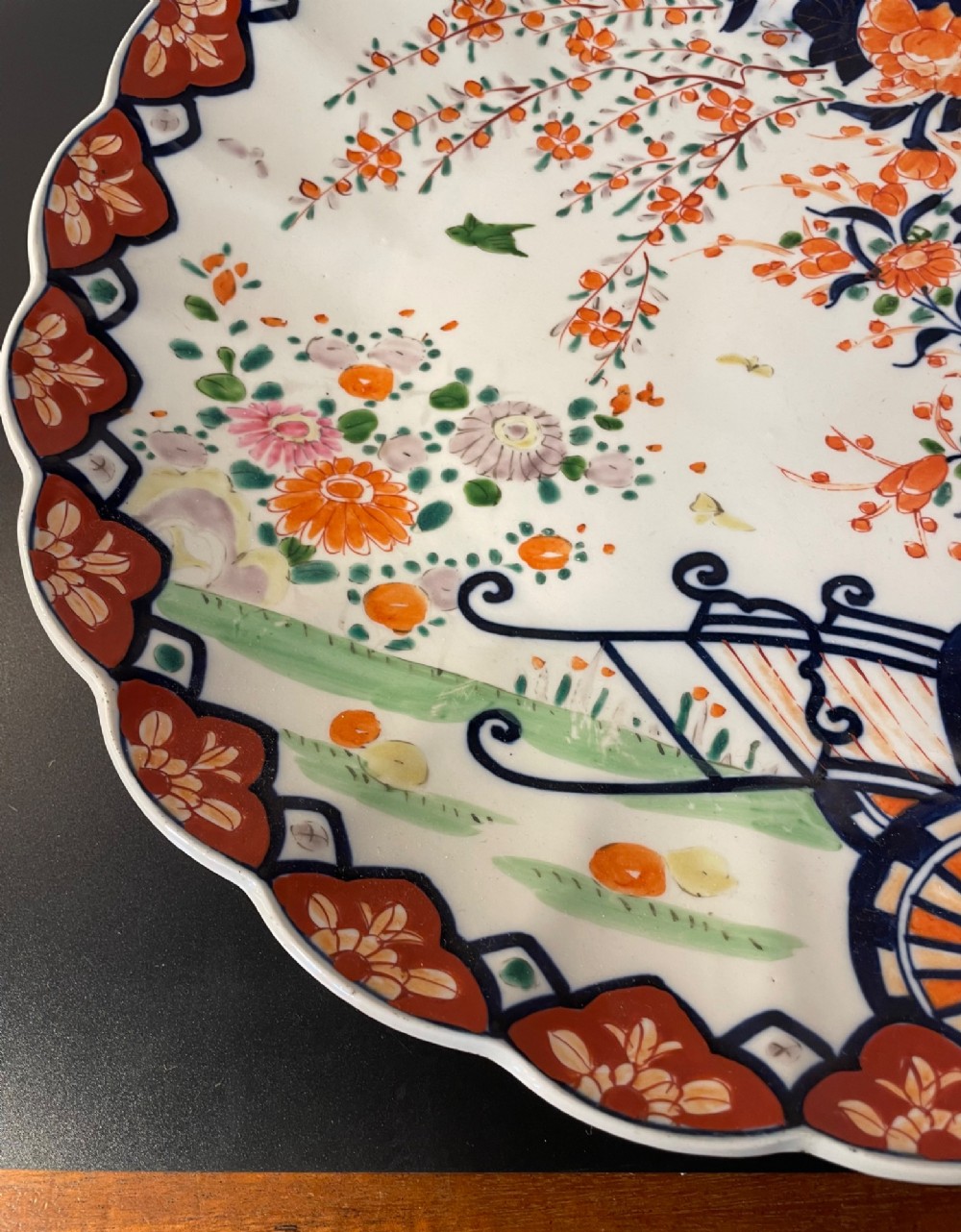 A circular Japanese Imari porcelain charger with a scalloped rim, featuring a hand-painted floral cart motif surrounded by cobalt blue and rust red enamel borders. View VI