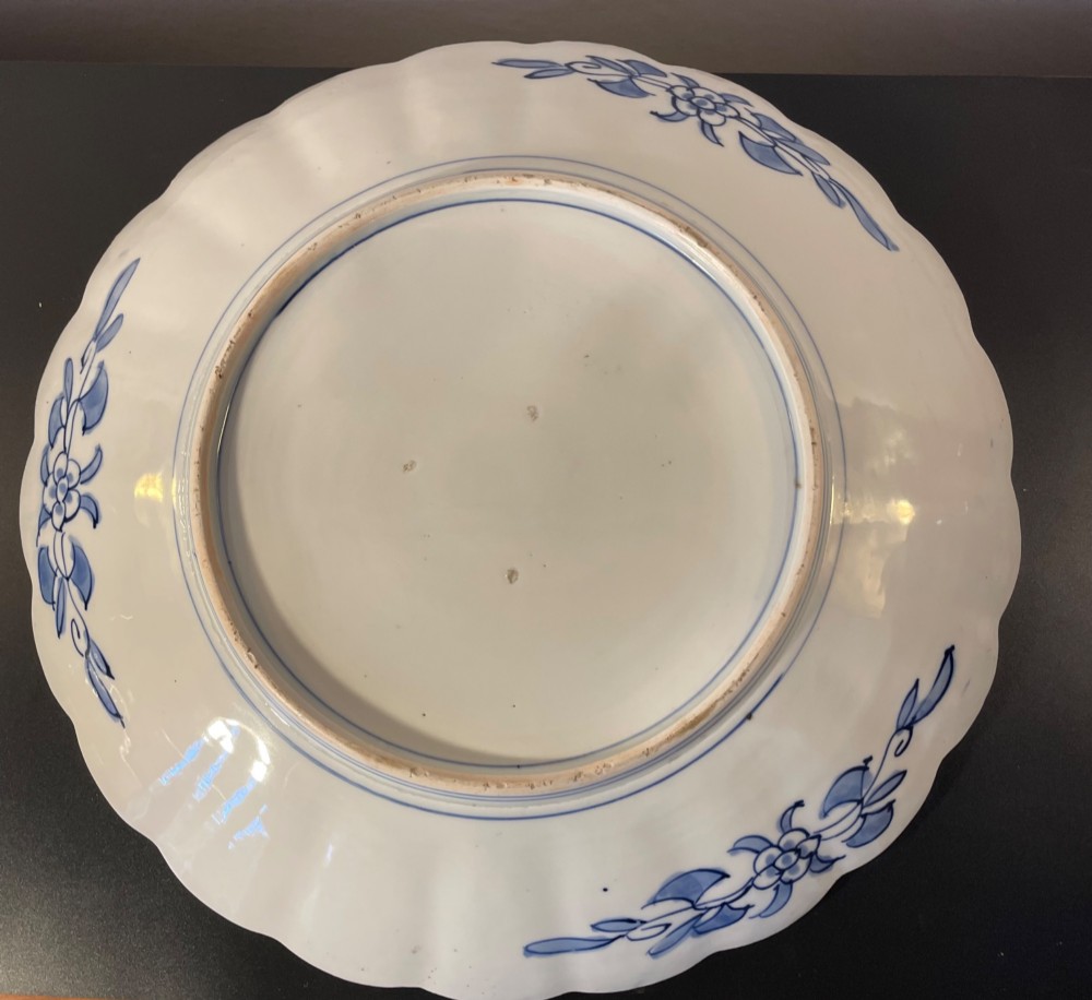 A circular Japanese Imari porcelain charger with a scalloped rim, featuring a hand-painted floral cart motif surrounded by cobalt blue and rust red enamel borders. View VII