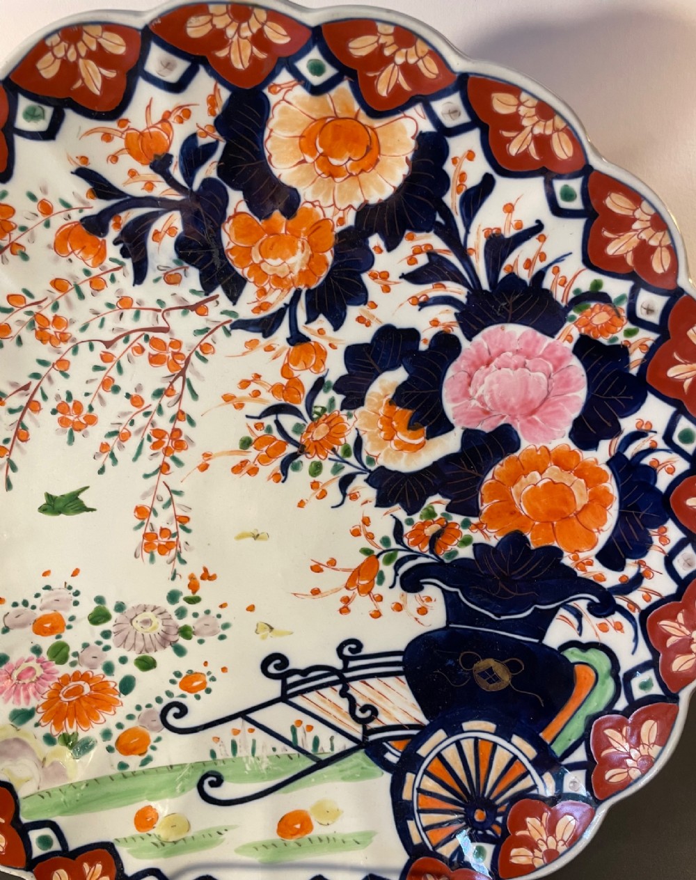 A circular Japanese Imari porcelain charger with a scalloped rim, featuring a hand-painted floral cart motif surrounded by cobalt blue and rust red enamel borders. View VIII