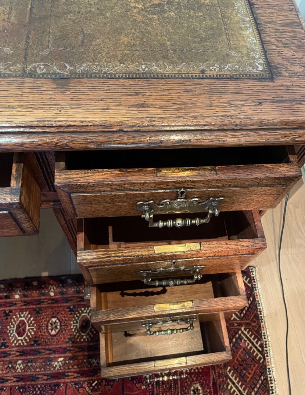 A Victorian oak pedestal desk featuring a gold-tooled leather writing top, nine drawers, and brass handles on a plinth base. View X