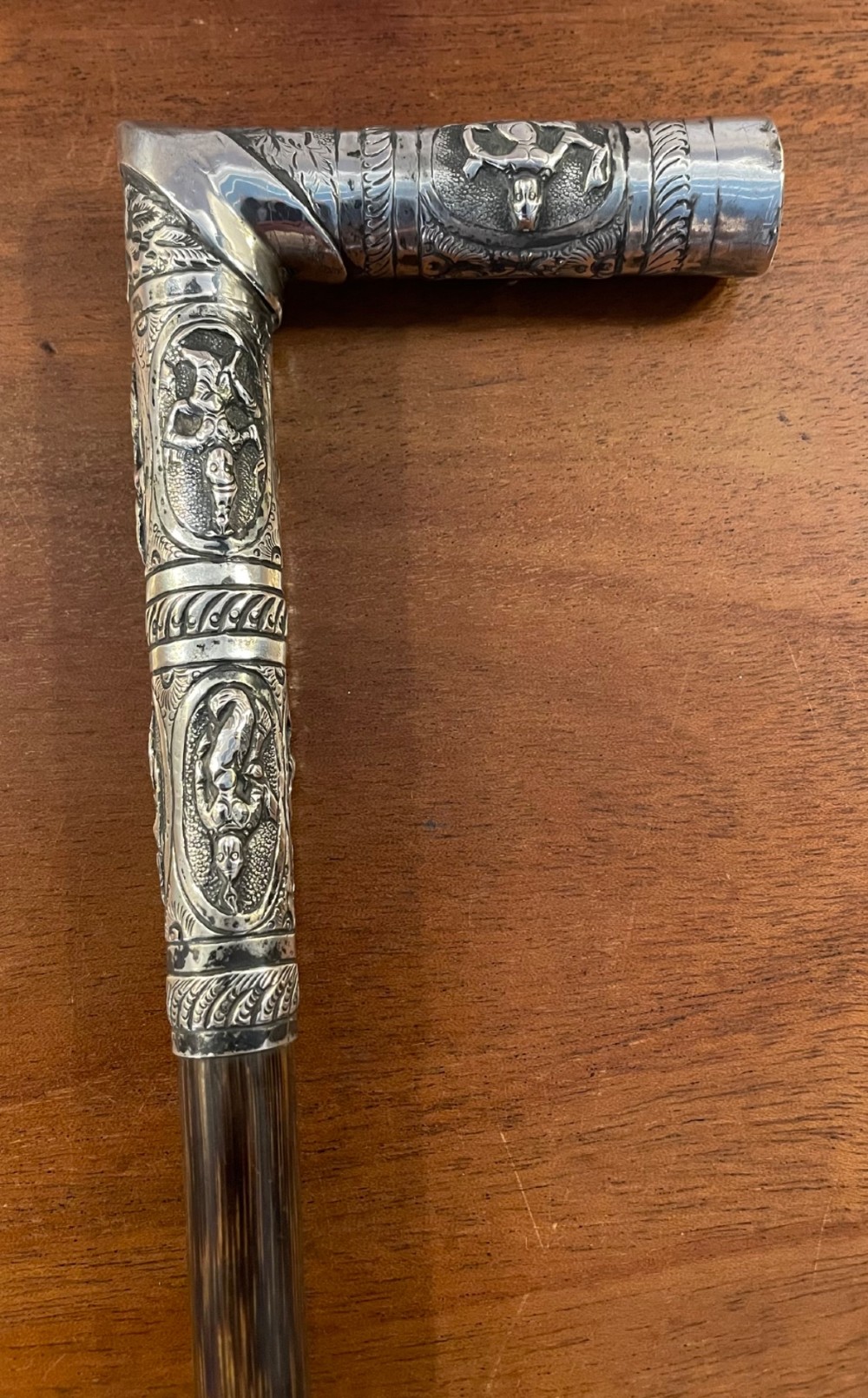 An antique walking stick with an embossed L-shaped silver handle featuring figurative motifs mounted on a dark brown bamboo shaft. View II