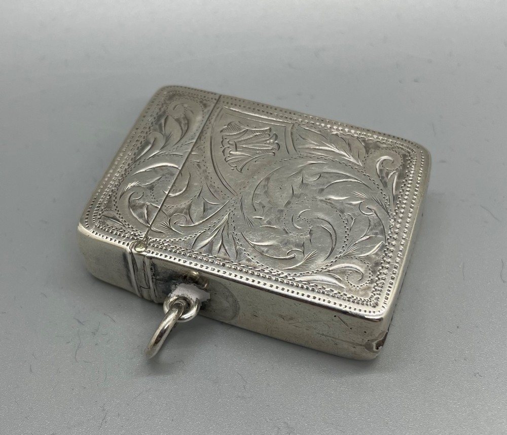 An upright rectangular sterling silver vesta case featuring hand-engraved foliate scrollwork, an unengraved cartouche, a hinged cover, and a side attachment ring. View II