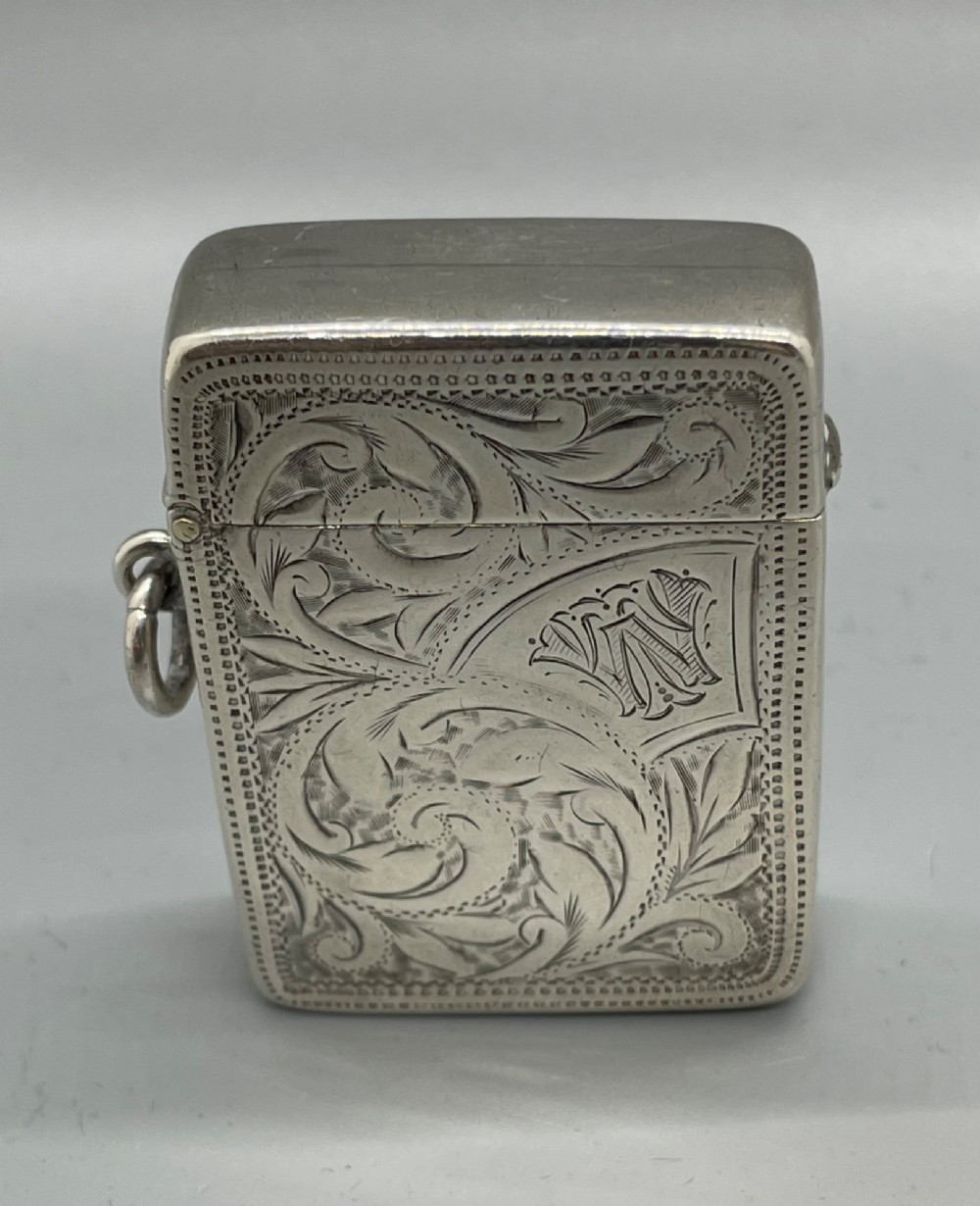 An upright rectangular sterling silver vesta case featuring hand-engraved foliate scrollwork, an unengraved cartouche, a hinged cover, and a side attachment ring. View III