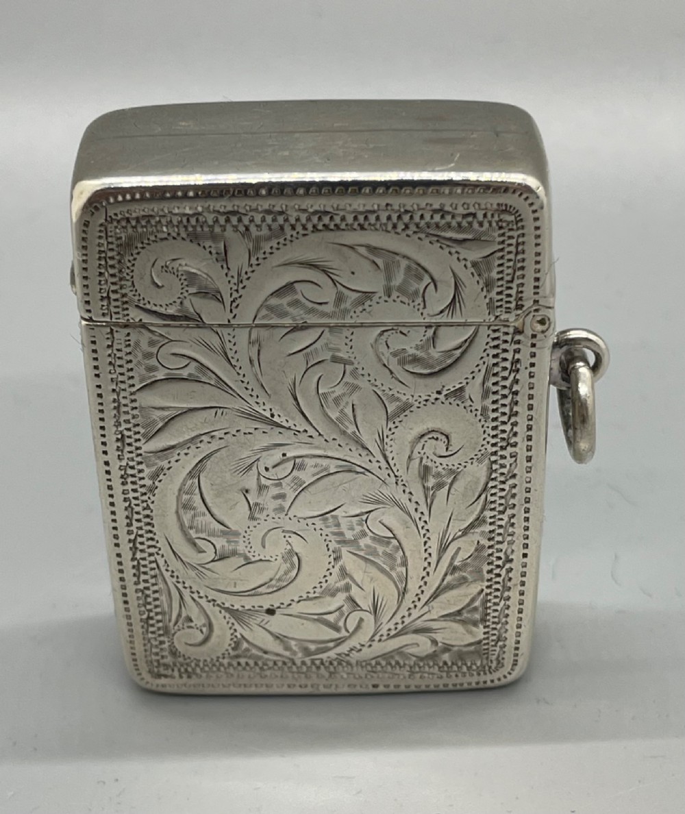 An upright rectangular sterling silver vesta case featuring hand-engraved foliate scrollwork, an unengraved cartouche, a hinged cover, and a side attachment ring. View IV