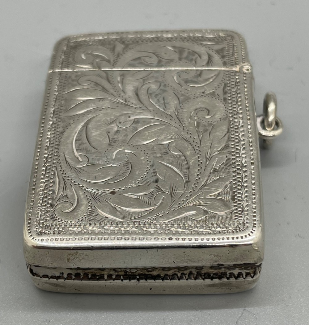 An upright rectangular sterling silver vesta case featuring hand-engraved foliate scrollwork, an unengraved cartouche, a hinged cover, and a side attachment ring. View V