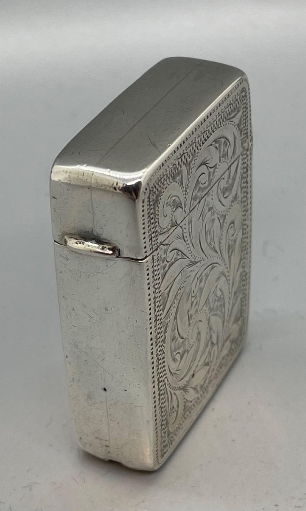 An upright rectangular sterling silver vesta case featuring hand-engraved foliate scrollwork, an unengraved cartouche, a hinged cover, and a side attachment ring. View VI