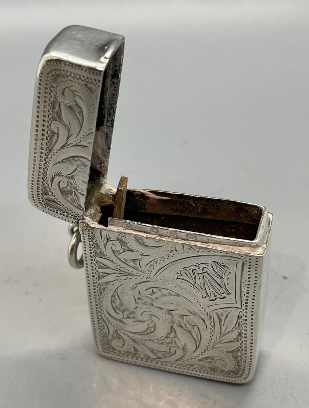 An upright rectangular sterling silver vesta case featuring hand-engraved foliate scrollwork, an unengraved cartouche, a hinged cover, and a side attachment ring. View VII