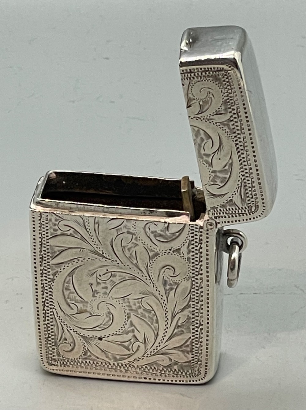 An upright rectangular sterling silver vesta case featuring hand-engraved foliate scrollwork, an unengraved cartouche, a hinged cover, and a side attachment ring. View IX