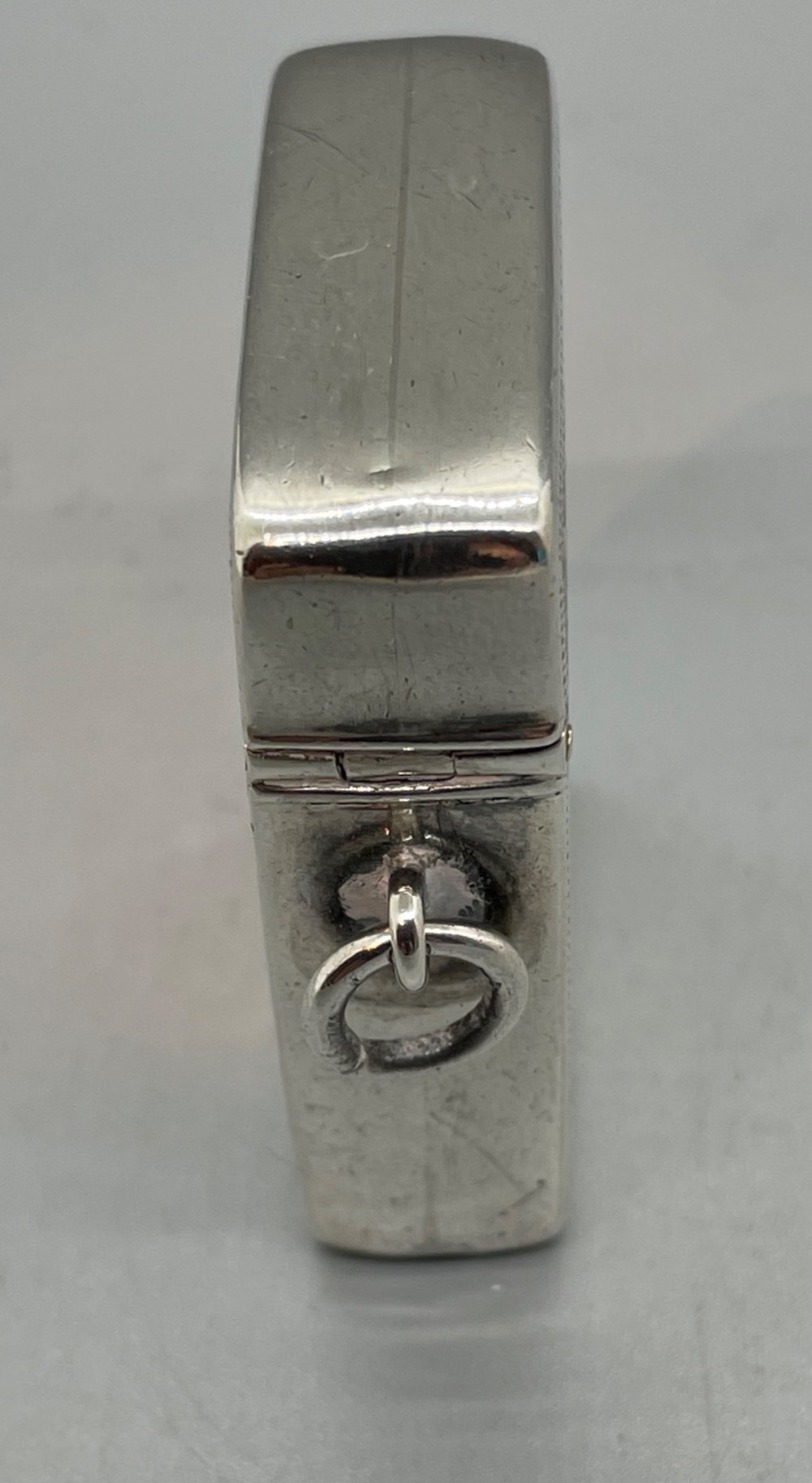 An upright rectangular sterling silver vesta case featuring hand-engraved foliate scrollwork, an unengraved cartouche, a hinged cover, and a side attachment ring. View X