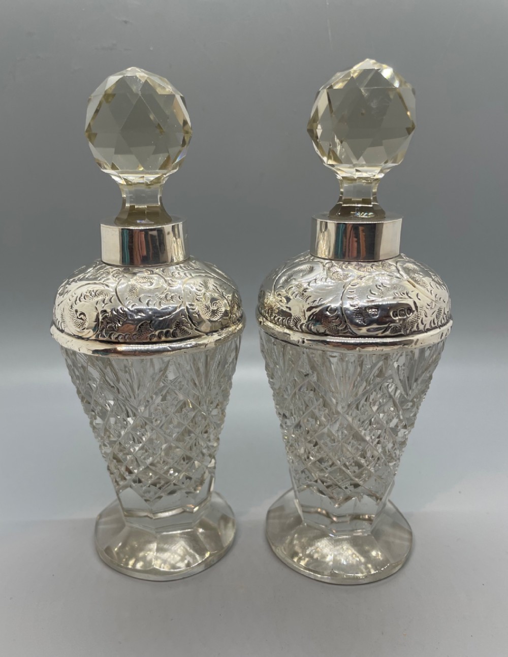 A pair of George V cut glass scent bottles featuring embossed sterling silver mounts and faceted glass stoppers.