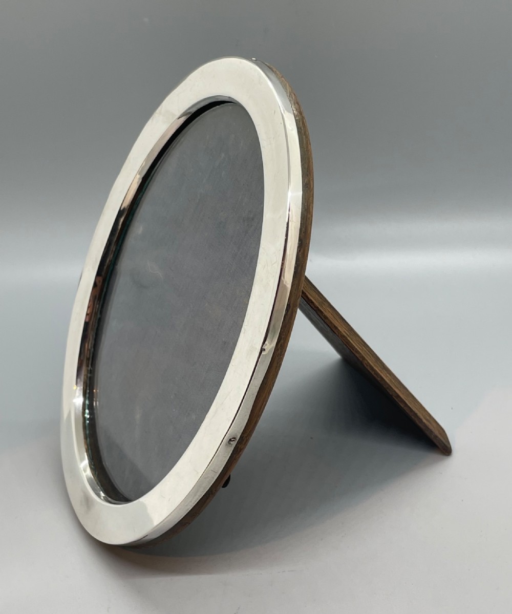 A George V oval sterling silver photo frame featuring a plain polished silver border and a dark wood easel backing. View III