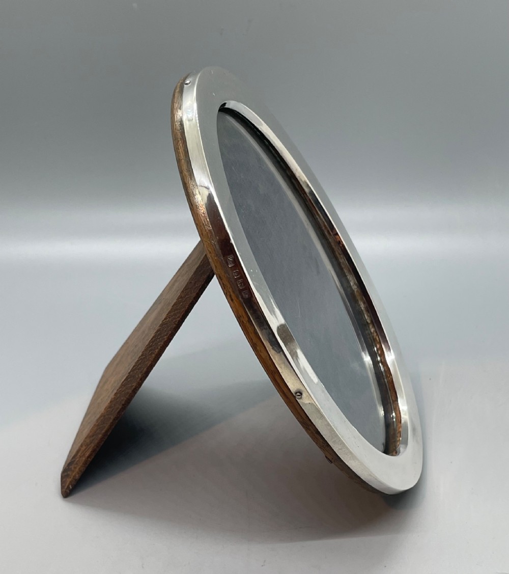 A George V oval sterling silver photo frame featuring a plain polished silver border and a dark wood easel backing. View IV