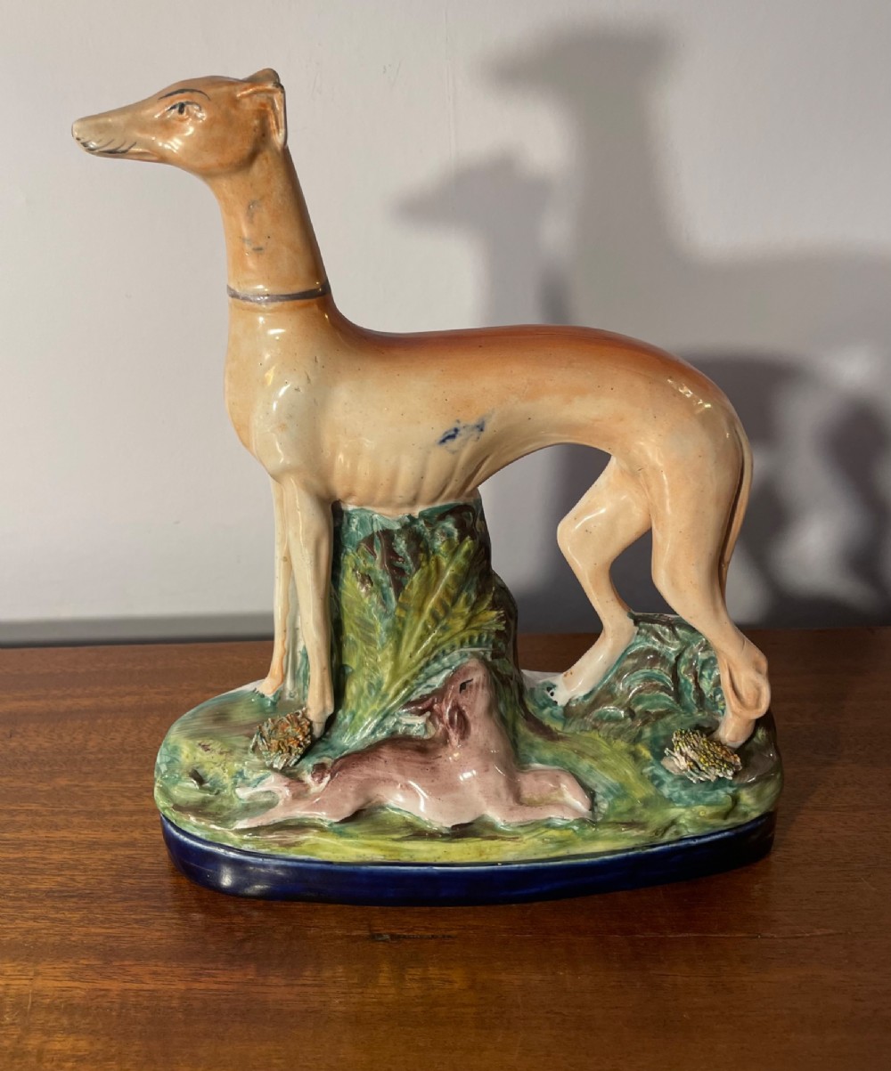 An antique Staffordshire earthenware figure depicting a standing tan hound with a crouching hare on a green and blue glazed base.