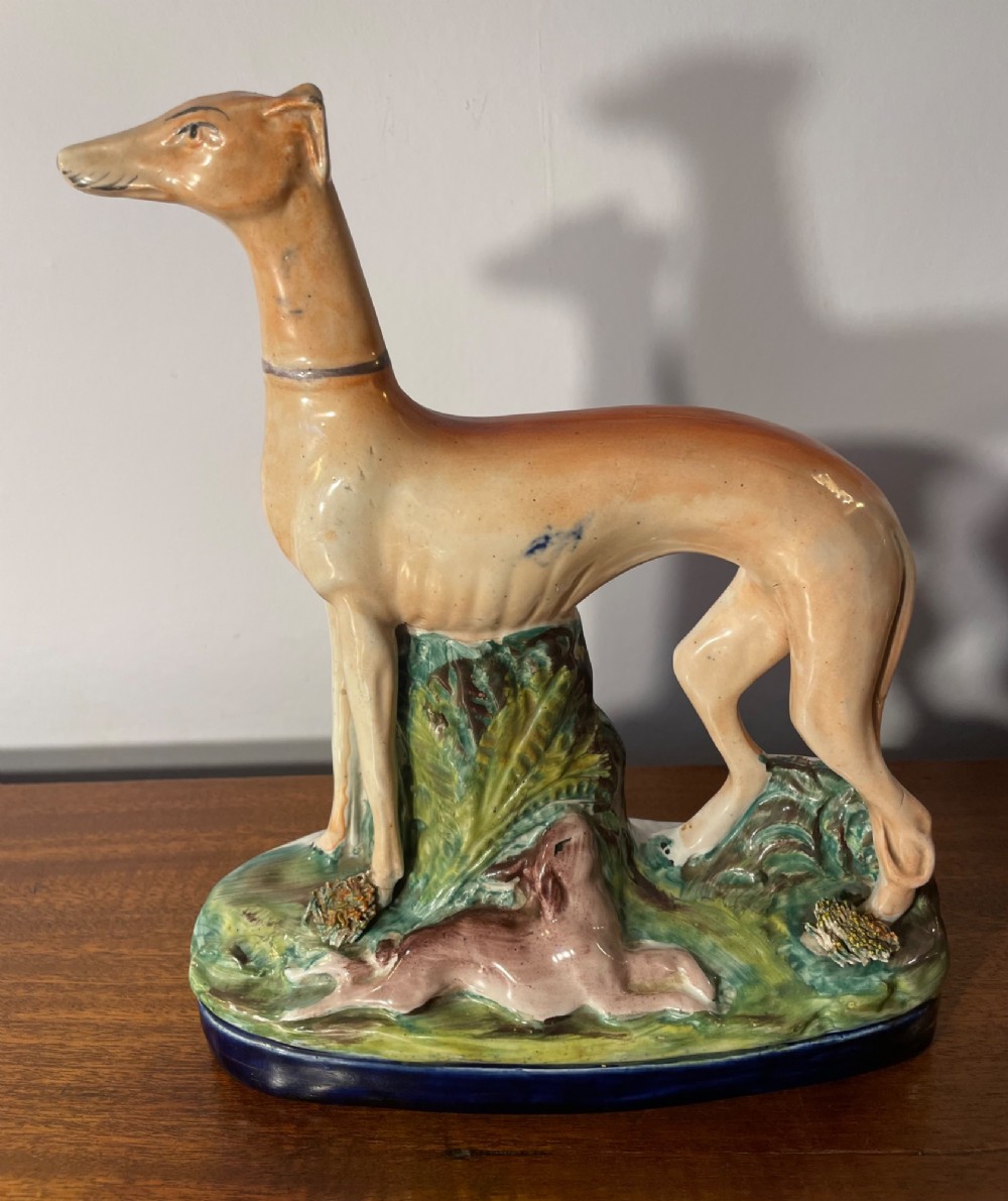An antique Staffordshire earthenware figure depicting a standing tan hound with a crouching hare on a green and blue glazed base. View II