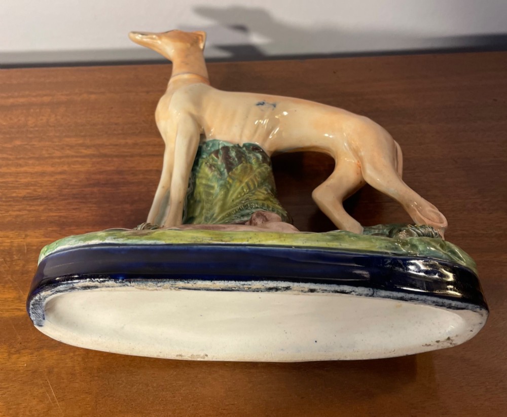 An antique Staffordshire earthenware figure depicting a standing tan hound with a crouching hare on a green and blue glazed base. View X