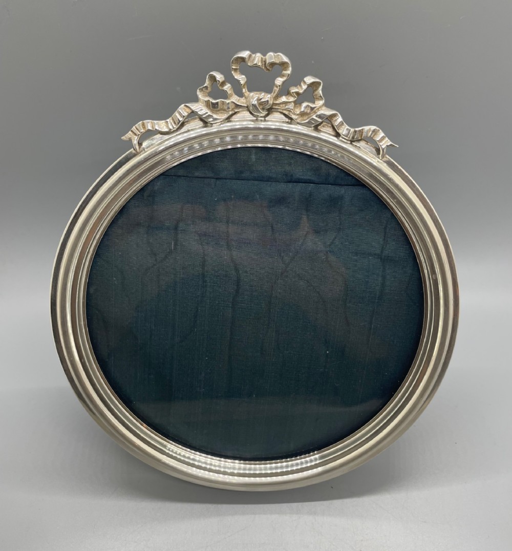 An oval sterling silver photo frame featuring a crest decorated with a ribbon bow, containing a dark fabric interior.