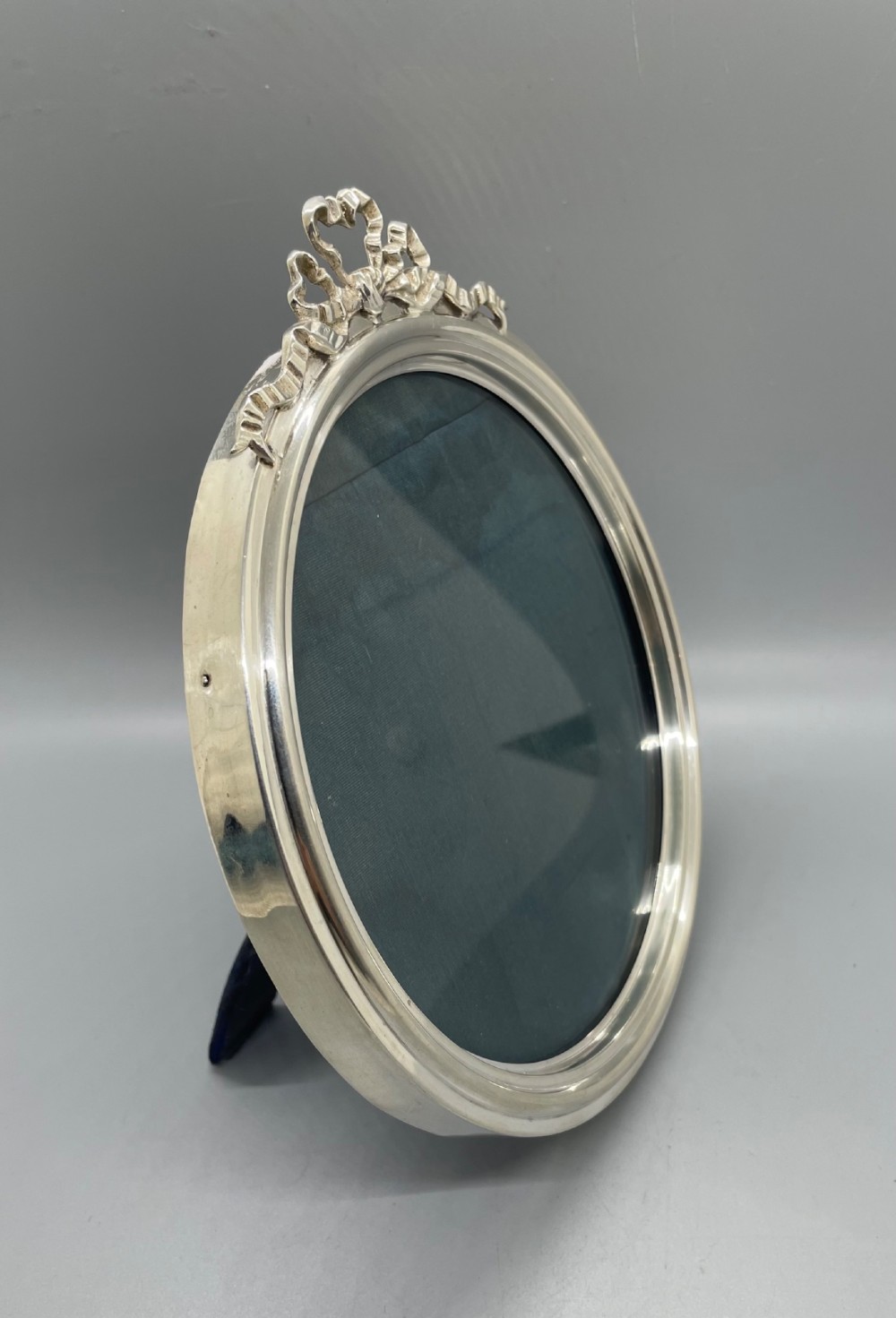 An oval sterling silver photo frame featuring a crest decorated with a ribbon bow, containing a dark fabric interior. View II