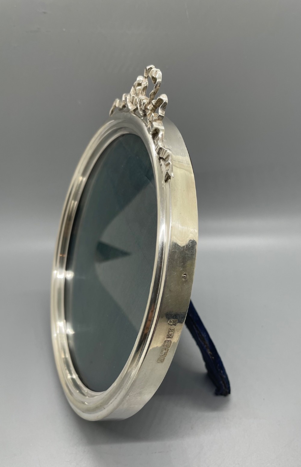 An oval sterling silver photo frame featuring a crest decorated with a ribbon bow, containing a dark fabric interior. View III