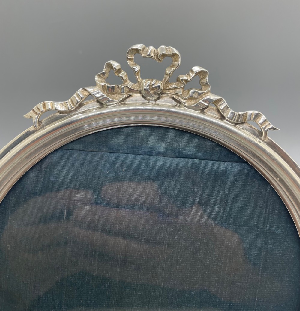 An oval sterling silver photo frame featuring a crest decorated with a ribbon bow, containing a dark fabric interior. View IV