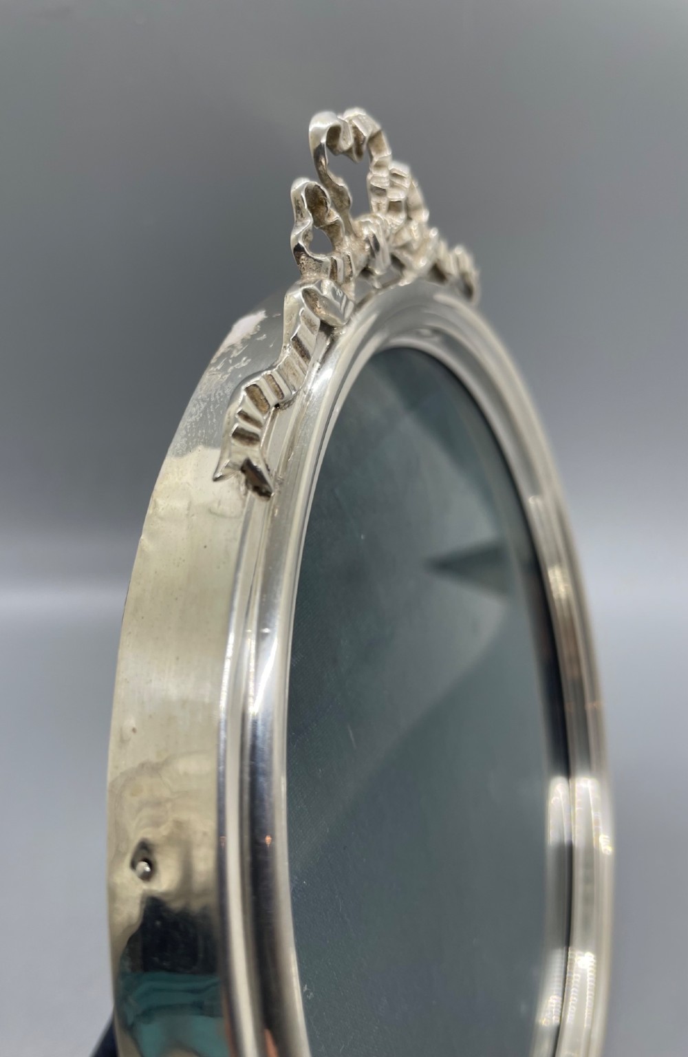 An oval sterling silver photo frame featuring a crest decorated with a ribbon bow, containing a dark fabric interior. View V