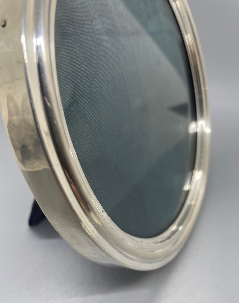 An oval sterling silver photo frame featuring a crest decorated with a ribbon bow, containing a dark fabric interior. View VI