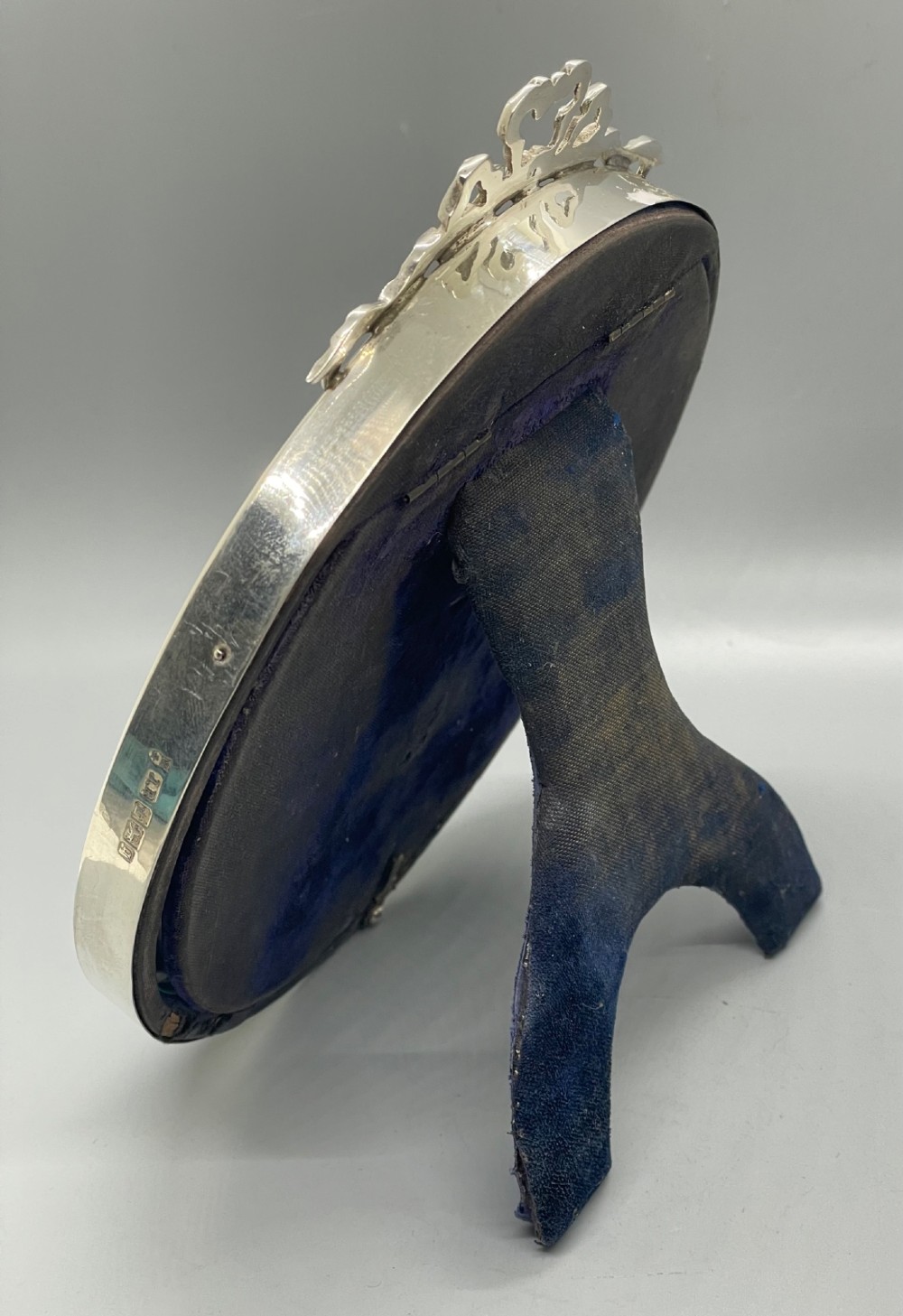 An oval sterling silver photo frame featuring a crest decorated with a ribbon bow, containing a dark fabric interior. View VIII