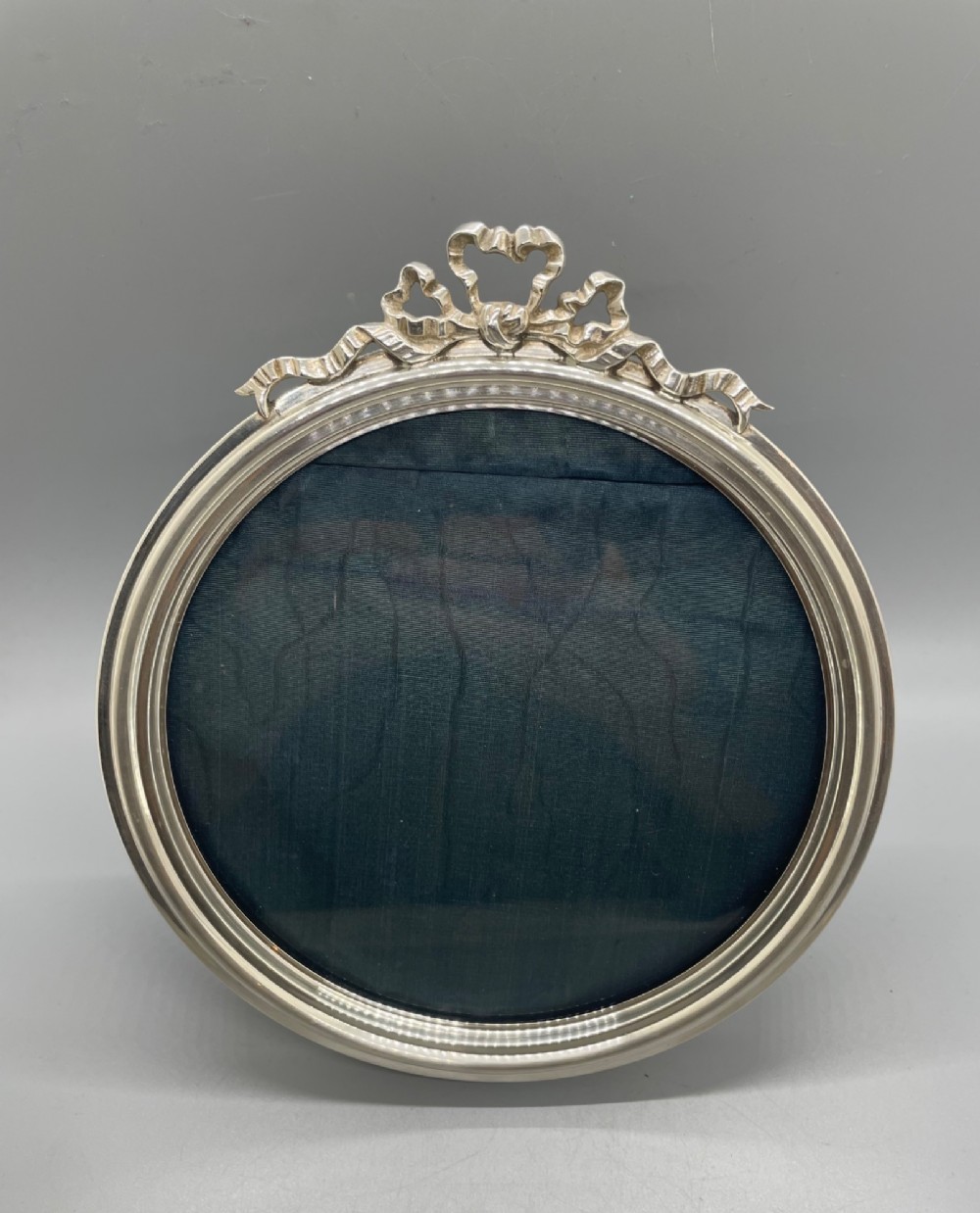 An oval sterling silver photo frame featuring a crest decorated with a ribbon bow, containing a dark fabric interior. View X