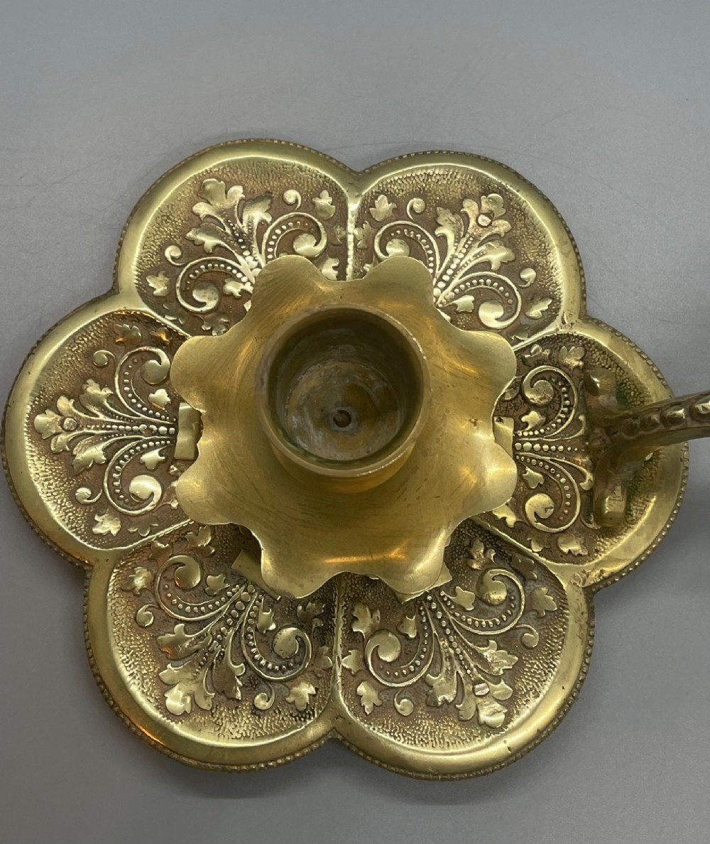 Late 19th-century French brass chamberstick featuring a scalloped petal tray with embossed scroll motifs, a central nozzle, and a cast ornate side handle. View VI